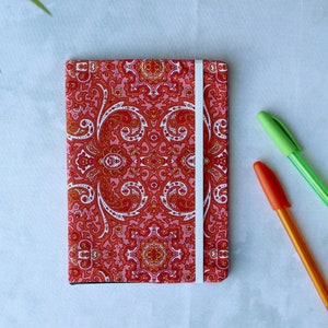 May include: A red and white patterned notebook with a white elastic closure. The notebook is lying on a white surface with three pens: one green, one blue, and one orange.