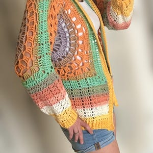 K&ouml;nnte beinhalten: Ein farbenfroher H&auml;kelcardigan mit einem Regenbogenstreifenmuster. Der Cardigan hat eine lockere Passform und eine lange L&auml;nge. Er ist aus verschiedenen Farben gefertigt, darunter Orange, Gelb, Gr&uuml;n und Pink.