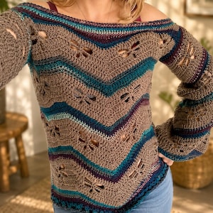 May include: A long-sleeved off-the-shoulder crochet sweater featuring a chevron pattern in shades of brown, teal, and dark purple. The garment is detailed with repeating dragonfly-shaped cutouts integrated into the knit design throughout the body and sleeves.
