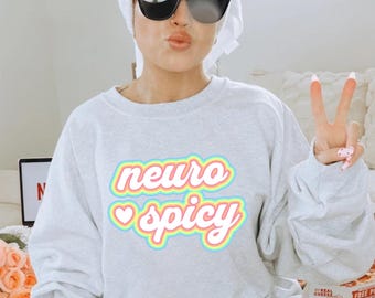 May include: A light gray sweatshirt with the text "neuro spicy" in a colorful, layered design. The sweatshirt has long sleeves and a crew neck. The text is outlined in multiple colors, including pink, yellow, and blue.