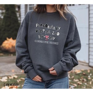 Pode incluir: Camisola cinza escuro com o texto branco "PERSONALLY VICTIMIZED BY MY CONNECTIVE TISSUES" e um coração rosa. A camisola é de estilo casual.