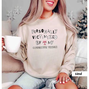 Op de afbeelding: Een licht beige sweatshirt met de tekst "PERSONALLY VICTIMIZED BY MY CONNECTIVE TISSUES" in zwarte letters. Een roze hart met een klein gezichtje staat tussen de woorden "BY" en "MY".