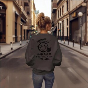 May include: A dark gray sweatshirt with a black smiley face with X's for eyes and a skeleton hand giving the middle finger. The text on the sweatshirt reads "Sometimes I just have to let God fix it. Cus if I fix it I'm going to jail."