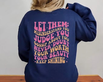 Op de afbeelding: Navyblauwe trui met een roze en oranje grafische print met de tekst "Let them: misunderstand you judge you gossip about you never doubt your worth your beauty keep shining".