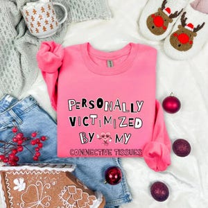Könnte beinhalten: Ein leuchtend pinkfarbenes Sweatshirt mit dem Text "PERSONALLY VICTIMIZED BY MY CONNECTIVE TISSUES" in Schwarz und Weiß. Das Bild enthält Jeansshorts, Weihnachtsschmuck, ein Lebkuchenhaus, eine Tasse und Rentier-Hausschuhe.