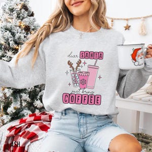 May include: A light grey sweatshirt featuring the text "dear SANTA just bring COFFEE" in pink. The graphic includes pink coffee cups and a glass with reindeer antlers. A white mug with Santa Claus is held in the hand.