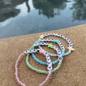 May include: Four beaded bracelets in shades of pink, green, and blue. The bracelets have white letter beads spelling out "TEAM", "BELLY", "CONRAD", and "COUSINS BEACH". Some bracelets feature star and seashell charms.
