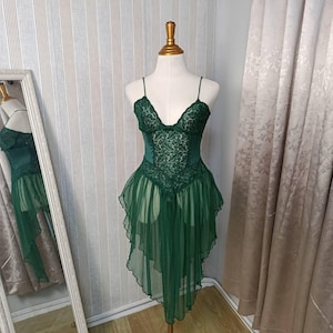May include: A dark green lingerie chemise featuring a lace bodice with thin spaghetti straps and a sheer, layered skirt with a high-low hemline. The garment is displayed on a mannequin, showcasing intricate floral lace detailing on the chest.