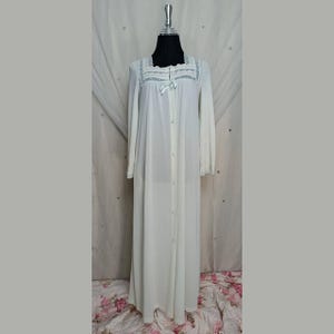 Puede incluir: Camisón largo blanco con mangas largas y botones en la parte delantera. El camisón presenta un escote cuadrado con ribete de encaje y un pequeño lazo. La prenda se exhibe en un maniquí.