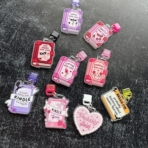 May include: A selection of acrylic keyrings in the shape of books and other designs. The keyrings feature various phrases such as "Fantasy Book Lover" and "Probably Smut." The keyrings have silver-colored hardware and are in various colors.