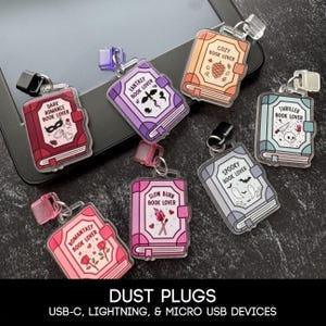 May include: A selection of book-shaped dust plugs in various colours, each themed with phrases like "Dark Romance" and "Fantasy Book Lover." The plugs have illustrations and text, with a clear plastic loop. The text "DUST PLUGS" is at the bottom of the image.