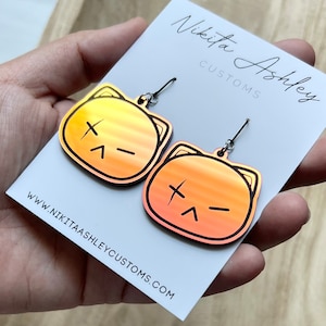 May include: A pair of cat-shaped earrings with a gradient of yellow to orange hues. Each earring features a black outline and a stylized face with an 'X' for an eye and a frown. The earrings are held on a white card with the text "Nikita Ashley CUSTOMS".