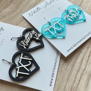 May include: Heart-shaped earrings in black and turquoise. The black earrings have white lettering that says "TXT" and "Huening Kai". The turquoise earrings have white lettering that says "TXT" and "Moa". The earrings are on white cards.