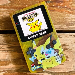 May include: A yellow Game Boy Color handheld video game console with a Pikachu design. The screen displays the Japanese text "Pocket Monsters" and a Pikachu character. The console has black buttons and a black directional pad.