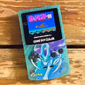 Op de afbeelding: Een turquoise Game Boy Color handheld-spelconsole met een blauwe en paarse Pokemon-personagegrafiek aan de voorkant. Het scherm toont de Japanse tekst "Pocket Monsters" en de gametitel "Pokemon Stadium".