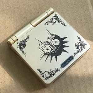 Puede incluir: Una Game Boy Advance SP plateada con un diseño en blanco y negro de Majora's Mask. El diseño presenta la máscara icónica con detalles intrincados y acentos florales. El logotipo de Nintendo es visible en la esquina inferior derecha.