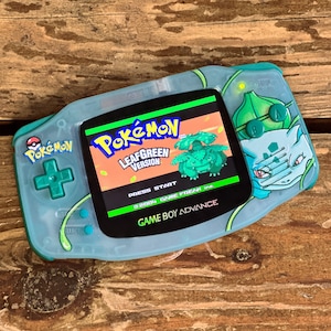 Peut inclure: Console portable Game Boy Advance de couleur turquoise affichant le jeu Pokémon LeafGreen Version. L'écran montre le titre du jeu et un graphique de Bulbasaur. La console présente un motif Bulbasaur sur le côté droit et le logo Pokémon.