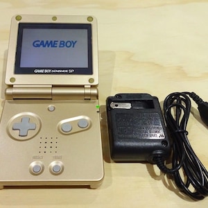 Puede incluir: Una consola de juegos portátil Game Boy Advance SP dorada con un adaptador de corriente y un cable negros. La consola tiene una pantalla que dice "GAME BOY" y "GAME BOY ADVANCE SP".