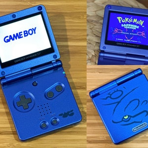 Puede incluir: Una Game Boy Advance SP azul con un logotipo "GAME BOY" en la pantalla. La consola está cerrada y la pantalla es visible. La consola tiene un diseño azul y negro con un botón de cruz negro y un botón de inicio negro. La consola está sobre una superficie de madera.