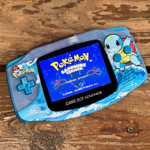 May include: A blue Game Boy Advance SP console with a custom design featuring a wave pattern and a Pokemon Sapphire game cartridge. The console has a blue and white screen with the text "POKÉMON SAPPHIRE VERSION" and "PRESS START".