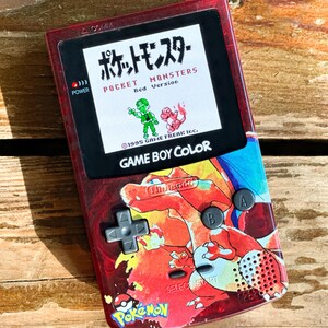 Könnte beinhalten: Eine rote Game Boy Color Handheld-Spielkonsole mit einem benutzerdefinierten Pokemon-Design. Die Konsole verfügt über ein rotes und orangefarbenes Charizard-Design mit einem schwarz-weißen Pokemon-Logo. Der Bildschirm zeigt den Titelbildschirm für Pokemon Red.