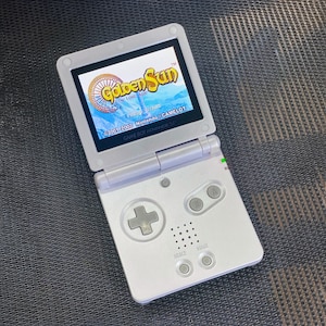 Puede incluir: Una consola de juegos portátil Game Boy Advance SP blanca, abierta para mostrar el juego "Golden Sun: The Lost Age". La pantalla muestra un gráfico colorido con texto. La consola tiene una cruceta, botones y el texto "GAME BOY ADVANCE SP".