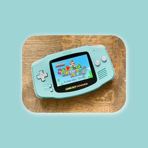 以下が含まれることがあります： ライトブルーのゲームボーイアドバンス携帯ゲーム機。白い十字キーとボタンが付いています。画面には「スーパーマリオアドバンス」のタイトル画面が表示されており、カラフルなグラフィックとテキストが映し出されています。デバイスには黒い画面ベゼルが付いています。