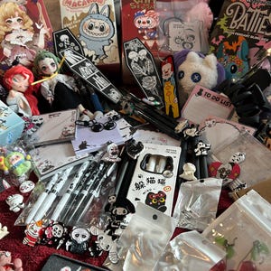 Puede incluir: Una colección de varios artículos, que incluyen peluches, bolígrafos, llaveros y pequeñas figuras. Los artículos presentan personajes de dibujos animados, calaveras y otros diseños de temática gótica. También hay cajas pequeñas y un bloc de notas con el texto "TO DO LIST".