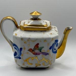 Könnte beinhalten: Eine weiße Porzellan-Teekanne mit goldenen Akzenten, einem goldenen Deckel und einem gebogenen Griff. Die Teekanne zeigt ein farbenfrohes Vogeldesign und blaue und goldene Blumenmuster. Ausguss und Griff sind ebenfalls goldfarben.