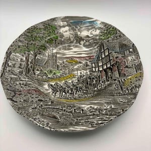 Può includere: Piatto decorativo con un disegno transferware in bianco e nero raffigurante una scena rurale con una carrozza trainata da cavalli, una chiesa e un ponte. Il piatto ha un bordo smerlato e una dettagliata illustrazione del paesaggio con tocchi di verde e giallo.