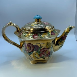 Può includere: Una teiera color oro con un motivo floreale. La teiera ha un manico e un beccuccio curvi, un coperchio con un pomello turchese ed è decorata con fiori colorati. La teiera è probabilmente in ceramica o porcellana.