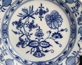 Op de afbeelding: Witte keramische plaat met een blauw bloemmotief. De plaat heeft een geschulpte rand en gedetailleerde blauwe ontwerpen van bloemen, bladeren en andere botanische elementen. Het ontwerp is gecentreerd en herhaald rond de rand.