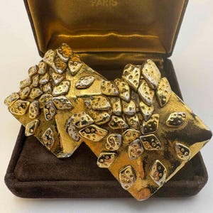 May include: A pair of vintage gold-tone earrings in a brown velvet box. The earrings are diamond-shaped with a textured surface and are adorned with small, irregular silver and gold-tone accents. The box is open, revealing the earrings.