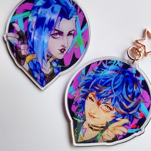 May include: Two acrylic keyrings featuring anime-style portraits with blue hair and colourful graffiti backgrounds. Each keyring has a star-shaped charm and a rose gold-coloured clasp. The portraits have a mischievous expression.