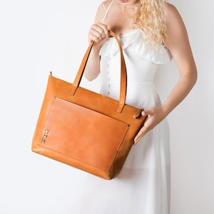 A girl holds a stylish honey-colored leather tote bag, a large light cognac purse with a spacious front pocket, made from genuine thick honey-colored leather.