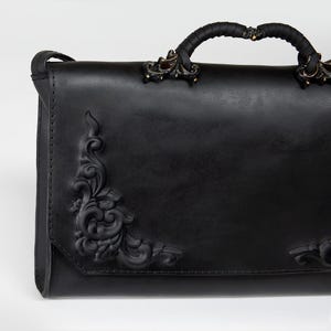 Könnte beinhalten: Eine schwarze Ledertasche mit verzierten Ecken und Griffdetails. Die Tasche hat ein erhabenes Blumendesign auf der Vorder- und den Seiten. Der Griff ist mit schwarzem Material umwickelt und mit bronzefarbenen Metallbeschlägen versehen.