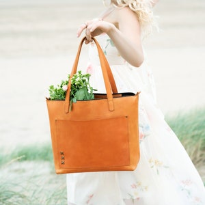 A girl holds a stylish honey-colored leather tote bag, a large light cognac purse with a spacious front pocket, made from genuine thick honey-colored leather.inside the bag there is a bouquet of flowers a good gift for a girl