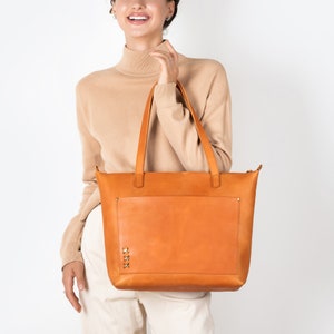 A girl holds a stylish honey-colored leather tote bag, a large light cognac purse with a spacious front pocket, made from genuine thick honey-colored leather.