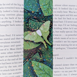 May include: A rectangular bookmark featuring a holographic finish with a dark green background of large leaves. A light green moth with white and red markings on its wings is centered on the bookmark, surrounded by small colorful star shapes.