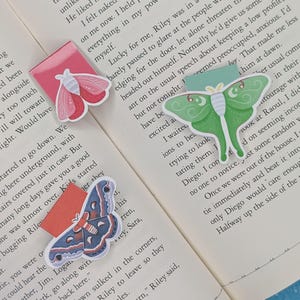 May include: Three decorative butterfly-shaped bookmarks. One is pink and red, another is green, and the third is blue and purple. Each bookmark has a rectangular tab. The bookmarks are on the pages of a book.