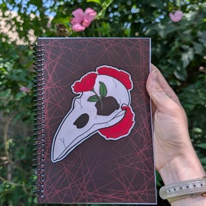 May include: A spiral-bound notebook with a dark brown cover featuring a red geometric line pattern. The center displays a graphic of a white bird skull with red carnations and a small green plant growing from the top of the skull structure.