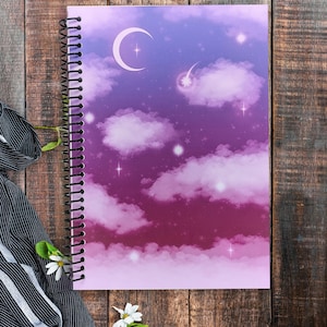 May include: A spiral-bound notebook featuring a night sky illustration with a gradient of purple and pink hues. The cover displays a crescent moon, a shooting star, and numerous small stars scattered among soft, fluffy clouds in shades of violet.