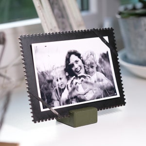 Può includere: Fotografia in bianco e nero incorniciata con una persona e due bambini che formano un cuore con le mani. La cornice ha un bordo decorativo ed è sostenuta da un piccolo supporto verde oliva.