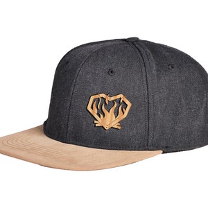 Könnte beinhalten: Dunkelgraue Baseballkappe mit flachem, beigefarbenem Wildleder-Schirm. Die Kappe hat ein Holz-Emblem in Herzform mit Flammen. Ein stilvolles Accessoire.