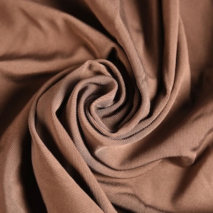 Könnte beinhalten: Nahaufnahme einer Wirbelung aus braunem Stoff. Das Material hat eine gerippte Textur und ist in weichen Falten drapiert. Die Farbe ist ein warmer, erdiger Ton mit subtilen Variationen von Licht und Schatten auf der Oberfläche.