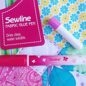 May include: Sewline Fabric Glue Pen with a pink and white design. The pen is on a patchwork fabric background. The packaging for the glue pen is pink and says "Sewline Fabric Glue Pen" and "Dries clear, water soluble."
