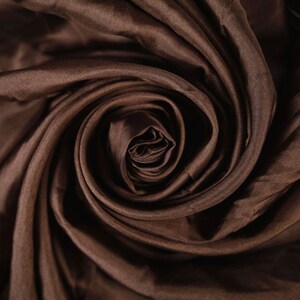 May include: Close-up of a swirl of brown fabric. The material is tightly gathered in a spiral, creating a textured, layered effect. The fabric has a subtle sheen, reflecting light and adding depth to the rich, dark brown color.