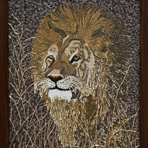 May include: A close-up of a lion's face, made of beads, with a brown and gold mane. The lion is looking directly at the viewer, with a serious expression. The background is a dark brown, with a few strands of gold beads representing grass.