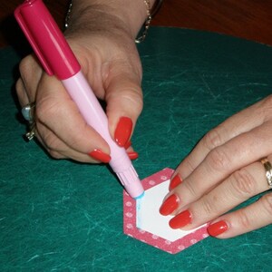 May include: A hand uses a pink marker to trace the outline of a white hexagon on a red and white polka dot paper hexagon.