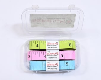 May include: Three colorful tape measures in a clear plastic case. The tape measures are green, pink, and blue. The green tape measure has the numbers 4 and 6 on it. The pink tape measure has the numbers 3, 4, and 5 on it. The blue tape measure has the numbers 3, 7, and 9 on it. Each tape measure has the text "The SewingHQ Where Happiness is Handmade Tape Measure" printed on it.